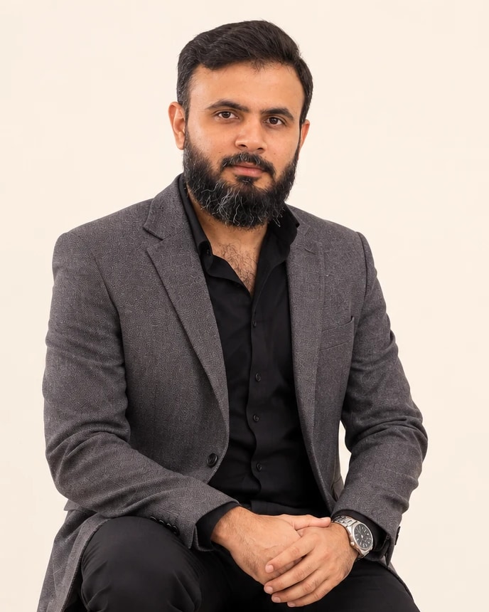 Portrait of Muhammad Hassan seated on a wooden stool in a grey blazer and black shirt